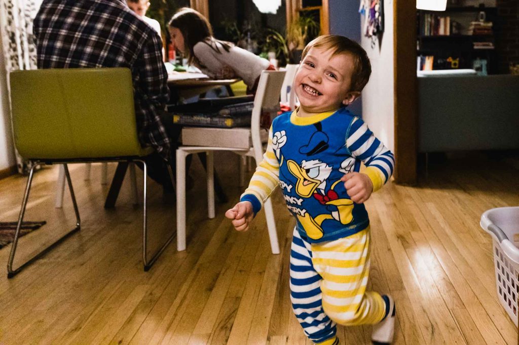 toddler in pajamas laughing and running through house while family sits at the table in the background